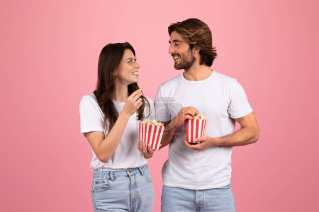 Photo for Happy young caucasian man and woman in white t-shirts eat popcorn at movie night, isolated on pink background, studio. Romantic date, food and snack, fun and entertainment together - Royalty Free Image