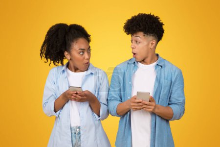 Photo for Astonished shocked African American man and woman in casual blue shirts look at each others smartphones, their faces expressing surprise and curiosity on a yellow backdrop - Royalty Free Image