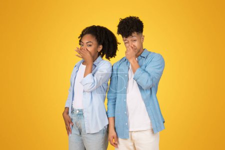 Photo for Two young black individuals in casual attire cover their mouths in a playful gesture, hinting at a secret or surprise, standing against a bright yellow backdrop with amused expressions - Royalty Free Image