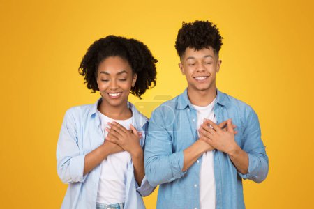 Téléchargez les photos : Une femme afro-américaine joyeuse et l'homme en vêtements décontractés tiennent leurs mains à leur cœur, souriant les yeux fermés, exsudant gratitude et joie sur un fond jaune, studio - en image libre de droit