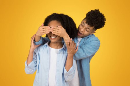 Photo for Happy young man in a denim shirt playfully covers a smiling womans eyes from behind, both dressed in light blue, sharing a moment of fun on a yellow background. Love, relationship, ad and offer - Royalty Free Image