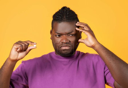 Photo for Portrait of unhappy black guy touching aching head and holding medical painkiller pill, suffering from discomfort and migraine pain, coping with unpleasant symptoms of hangover on yellow background - Royalty Free Image