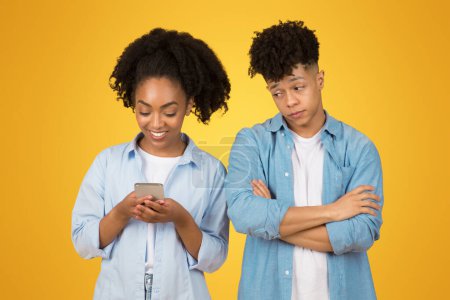 Foto de Una joven afroamericana sonriente está absorta en su teléfono inteligente mientras un hombre joven está a su lado con los brazos cruzados, mirando ligeramente molesto, sobre un fondo amarillo - Imagen libre de derechos