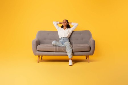 Photo for Happy carefree African American teen girl relaxing sitting on sofa over yellow studio background. Youngster lady enjoying rest holding hands behind head. Full length shot - Royalty Free Image