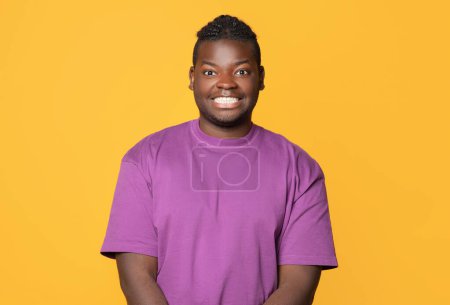 Foto de Tipo negro dentado posando en púrpura camiseta sonriendo a la cámara sobre fondo amarillo, Studio Shot. Retrato de un joven afro-americano positivo expresando emociones positivas - Imagen libre de derechos