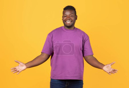 Téléchargez les photos : Gai noir guy dans pourpre t-shirt écartant les bras sourire à caméra debout sur jaune Studio toile de fond. Positif Afro-Américain Millénaire Homme Posant Exprimant Positivité - en image libre de droit