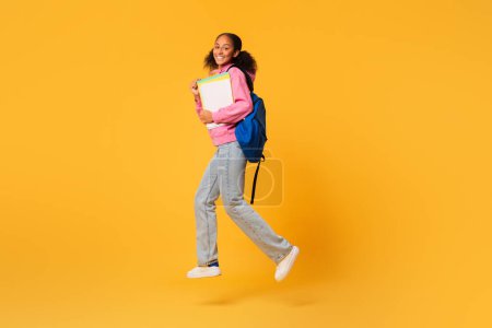 Téléchargez les photos : Heureuse adolescente noire sautant avec un sac à dos bleu et des cahiers d'exercices universitaires sur fond de studio jaune. Joie de la formation académique, concept apprenant réussi. Longueur totale - en image libre de droit