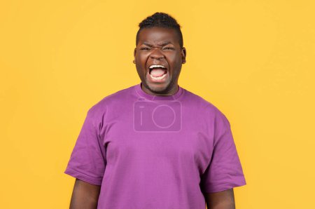 Téléchargez les photos : Portrait de l'homme afro-américain en colère en t-shirt violet criant fort exprimant des émotions négatives, posant sur fond de studio jaune, criant sur les problèmes ou les besoins - en image libre de droit