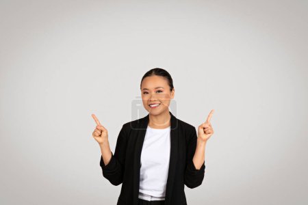 Téléchargez les photos : Femme d'affaires asiatique joyeuse dans un costume noir pointant vers le haut avec les deux mains, souriant et regardant vers de nouvelles opportunités ou présentant quelque chose au-dessus, sur un fond gris - en image libre de droit