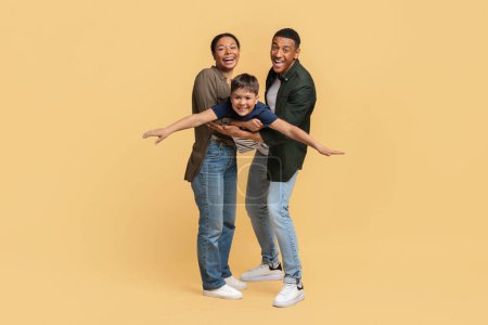 Photo for Happy family of three father, mother and teen aged son having fun on colorful studio background. Cheerful young african american parents lifting their smiling kid boy imitating airplane, full length - Royalty Free Image