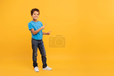 Téléchargez les photos : Joyeux garçon noir aux cheveux bouclés pointant latéralement vers l'espace libre et souriant à la caméra, portant un t-shirt bleu et un jean sur fond jaune uni, pleine longueur - en image libre de droit