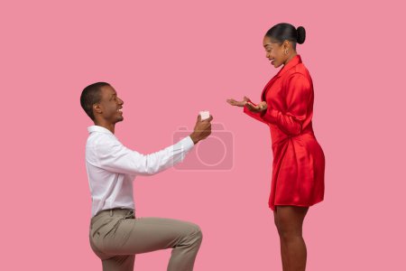 Photo for Smiling black man on one knee holding open ring box, proposing to overjoyed woman in red dress, both captured in moment of engagement bliss against soft pink backdrop - Royalty Free Image