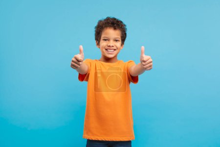 Photo for Cheerful curly-haired boy in orange t-shirt giving two enthusiastic thumbs up, showing positivity and approval against blue backdrop - Royalty Free Image