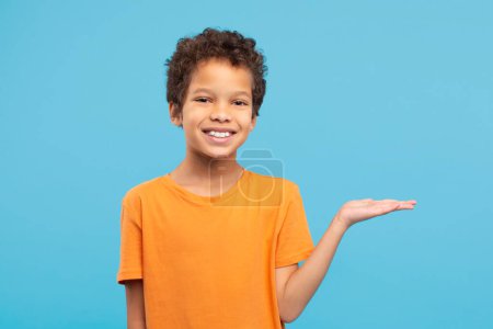 Photo for Smiling boy with curly hair, wearing orange t-shirt, extends his hand in presenting gesture, showcasing invisible product or idea on blue backdrop - Royalty Free Image