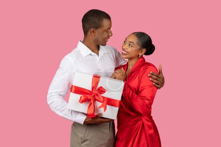 Téléchargez les photos : Délicieux homme noir en chemise blanche présente cadeau avec ruban rouge à la femme reconnaissante en robe rouge, partageant moment joyeux de donner sur fond rose - en image libre de droit