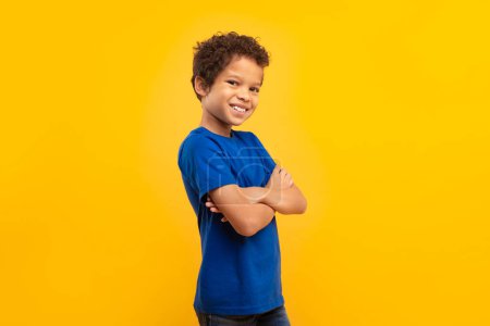 Photo for Self-assured latin boy with curly hair and broad smile, arms crossed wearing blue t-shirt, standing against vivid yellow background - Royalty Free Image