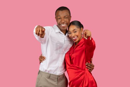 Téléchargez les photos : Couple noir engageant pointant directement vers la caméra avec des sourires confiants, homme vêtu d'une chemise blanche et femme en robe rouge, sur fond rose doux - en image libre de droit