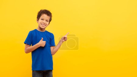 Foto de Alegre niño de pelo rizado en camiseta azul dando pulgares hacia arriba y apuntando hacia un lado en el espacio de copia mientras sonríe, posando sobre un fondo amarillo soleado, panorama - Imagen libre de derechos