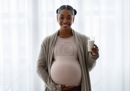 Photo for Cheerful african american expecting woman holding glass of milk, kefir or yogurt. Pregnant black lady enjoying dairy products during pregnancy, standing next to window at home - Royalty Free Image