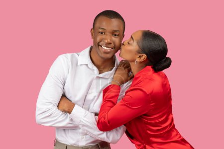 Foto de Elegante mujer negra en vestido rojo cariñosamente besando mejilla de hombre sonriente en camisa blanca, expresando amor y conexión contra telón de fondo rosa - Imagen libre de derechos