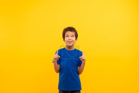 Photo for Curly-haired boy in blue t-shirt looking and pointing upwards at free space with look of curiosity set against bright yellow background, banner - Royalty Free Image