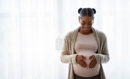 Foto de Sonriente joven embarazada negra con ropa cómoda cogida de la mano sobre su gran vientre, disfrutando de su embarazo, esperando un bebé, de pie junto a la ventana en casa, panorama con espacio para copiar - Imagen libre de derechos