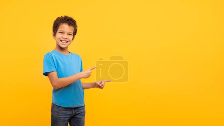 Téléchargez les photos : Joyeux garçon latin aux cheveux bouclés en chemise bleue pointant vers son côté à l'espace libre avec un sourire joyeux, sur fond jaune vif, panorama - en image libre de droit