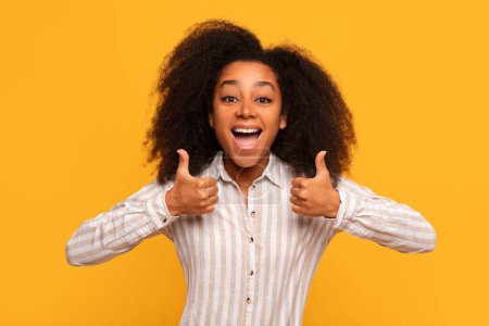 Téléchargez les photos : Jeune femme noire exubérante aux cheveux bouclés moelleux donne avec enthousiasme deux pouces vers le haut, ses yeux scintillant de joie, sur fond jaune monochrome - en image libre de droit