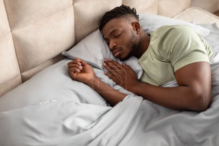 Téléchargez les photos : Un sommeil sain. Angle élevé de l'homme afro-américain couché confortablement endormi dans un lit confortable dans la chambre à coucher de la maison. Jeune homme au repos les yeux fermés à l'intérieur. Relaxation domestique et temps de sieste concept - en image libre de droit