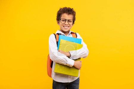 Foto de Niño nerd sonriente con el pelo rizado y gafas, agarrando los libros de la escuela y con una mochila naranja, listo para la clase contra el fondo amarillo - Imagen libre de derechos