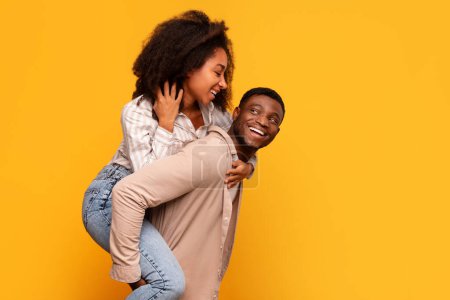 Photo for Happy black couple enjoying playful moment with man giving piggyback ride to laughing woman, both radiating joy against yellow backdrop, free space - Royalty Free Image
