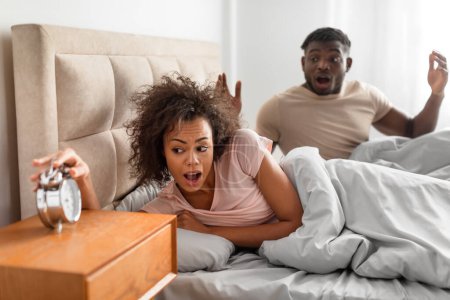 Photo for Shocked funny young african american spouses overslept looking at alarm clock in bed, being late waking up in panic after night sleep in modern bedroom at home. Late for work, oversleeping - Royalty Free Image