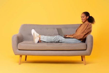 Foto de Chica adolescente afroamericana relajándose con su computadora portátil viendo películas en línea mientras está sentada en el sofá contra el fondo amarillo del estudio. Estudiante dama disfruta de internet de ocio y entretenimiento - Imagen libre de derechos