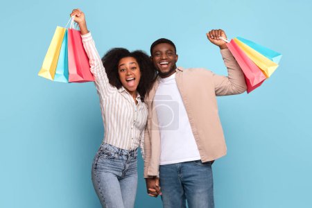 Téléchargez les photos : Joyeux jeune couple noir célèbre leur frénésie de shopping, soulevant joyeusement leurs sacs colorés sur fond bleu vibrant - en image libre de droit