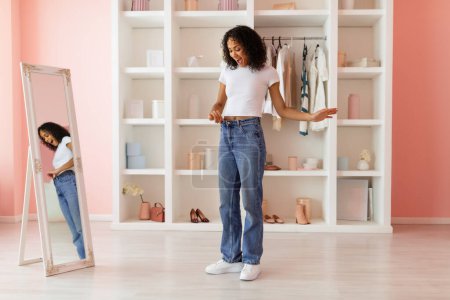 Foto de Mujer joven asombrada mostrando su pérdida de peso mediante la colocación en los pantalones vaqueros grandes, de pie en una habitación bien iluminada con elegante interior blanco y rosa - Imagen libre de derechos