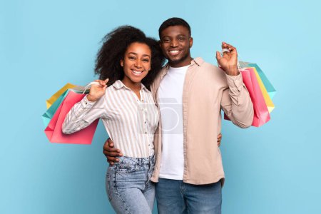 Photo for Smiling young black couple enjoys successful shopping spree, holding colorful bags, radiating satisfaction and happiness on refreshing light blue background - Royalty Free Image