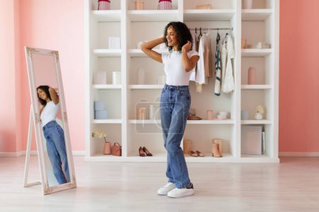 Téléchargez les photos : Jeune femme rayonnante aux cheveux bouclés essayant un jean taille haute, admirant son reflet dans un miroir dans une chambre rose chic, prise de vue pleine longueur, espace libre - en image libre de droit