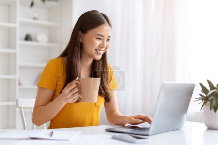 Photo for Distance Work. Beautiful Asian Female Freelancer Drinking Coffee And Working With Laptop While Sitting At Desk In Home Office, Smiling Young Korean Woman Typing On Computer Keyboard, Closeup - Royalty Free Image