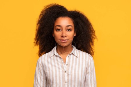 Photo for Calm young woman with gentle smile and voluminous curly hair wears neutral striped shirt, posing against smooth yellow background - Royalty Free Image