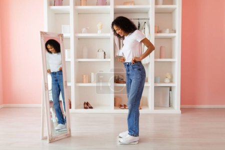 Photo for Young latin woman in casual attire happily admiring her fit in blue jeans and white t-shirt, reflected in full-length mirror in a stylish room - Royalty Free Image