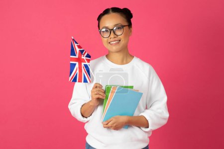 Photo for Beautiful young asian student woman holding flag of United Kingdom and books, happy korean female recommending international studies and exchange programs, standing against bright pink background - Royalty Free Image