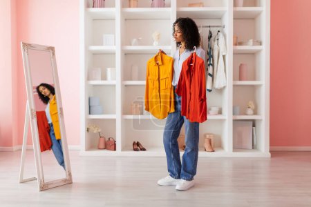 Téléchargez les photos : Femme indécise tenant des chemises jaunes et rouges, contemplant son choix de tenue dans un miroir dans une pièce aux murs roses et aux étagères blanches - en image libre de droit