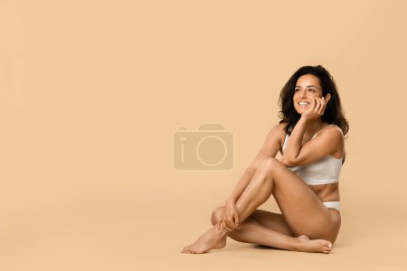 Téléchargez les photos : Belle femme en sous-vêtements blancs assise sur le sol et regardant ailleurs, jolie femme souriante qui rit joyeusement avec sa main sur sa joue, posant sur fond de studio beige, espace de copie - en image libre de droit
