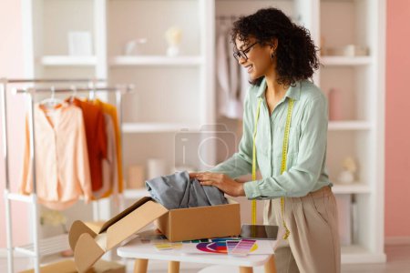 Photo for Smiling female fashion designer unpacking new garment from cardboard box, preparing for creative session in her pastel-themed studio - Royalty Free Image