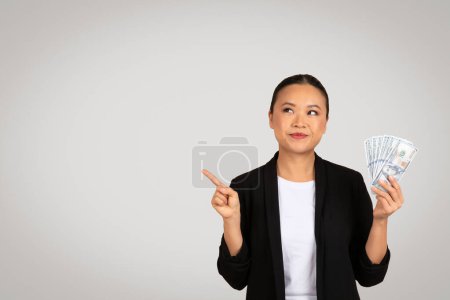 Téléchargez les photos : Femme d'affaires asiatique intelligente tenant un fan de billets en dollars américains et pointant du doigt, indiquant peut-être des opportunités d'investissement rentables ou un succès financier, dans un contexte neutre - en image libre de droit