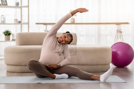 Téléchargez les photos : Joyeux jeune femme noire enceinte faisant de l'exercice à la maison, assise sur un tapis de yoga dans le salon, étirant son corps et souriant. Fitness pour les femmes enceintes, mode de vie sain - en image libre de droit