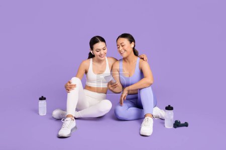 Photo for Two diverse women in sportswear sitting on floor, looking at smartphone together, enjoying break with water bottles and dumbbell nearby on purple background - Royalty Free Image