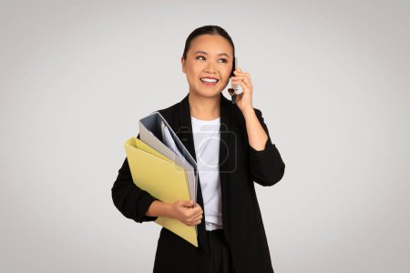 Photo for Engaged Asian businesswoman in a black blazer talking on the phone while holding colorful folders, portraying active business communication and multitasking, against a light grey backdrop - Royalty Free Image