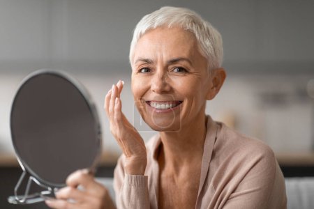 Foto de Retrato de una atractiva anciana sosteniendo el espejo sonriendo a la cámara mientras aplica humectante facial y cuida la piel de la cara. Prevención de arrugas, Cuidado de la piel para el envejecimiento de la piel - Imagen libre de derechos