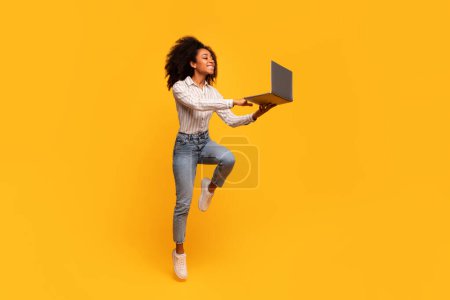 Photo for Energetic young black woman with curly hair joyfully jumps in mid-air, holding out her laptop as if in dynamic presentation, against vivid yellow backdrop - Royalty Free Image